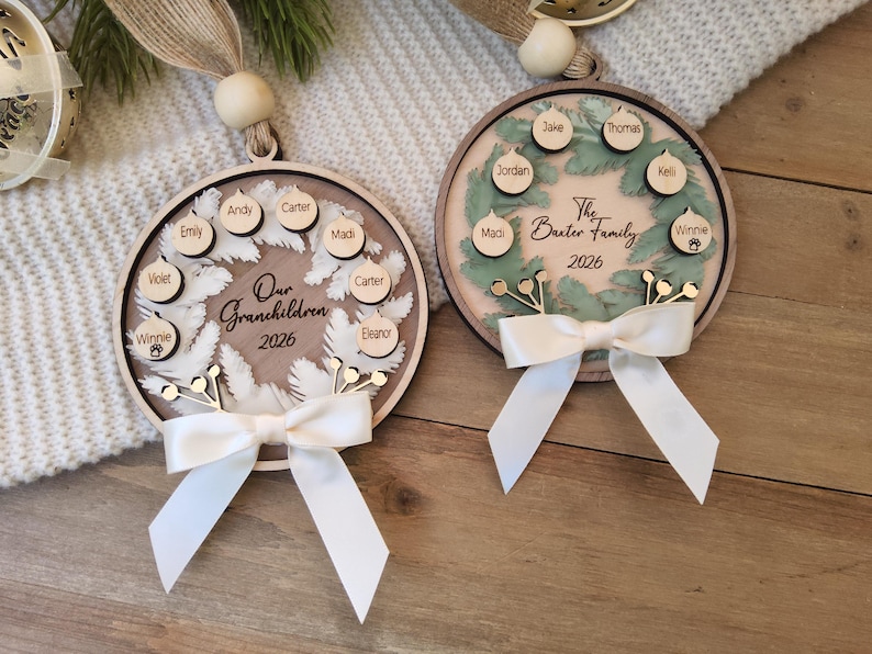 May include: Two circular wooden ornaments featuring a wreath design, white ribbon bows, and hanging loops with wooden beads. One ornament displays the text Our Grandchildren 2026 with names on small wooden circles. The other shows The Baxter Family 2026.