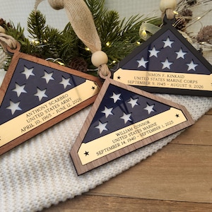 May include: Three triangular wooden ornaments featuring a dark blue inlay with cut-out stars and a gold-toned metal plaque. Each plaque is engraved with a name, military branch, and dates. The ornaments hang from tan ribbons with wooden beads.