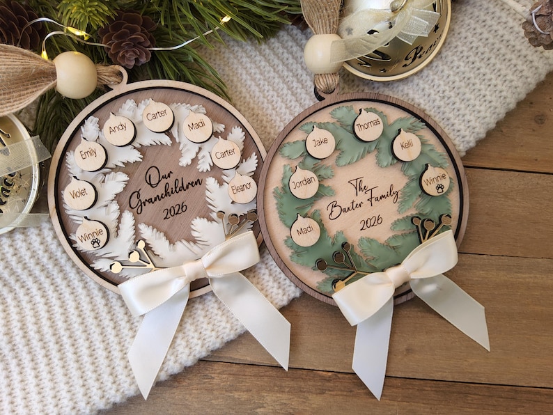 May include: Two circular wooden ornaments featuring a wreath design. The left ornament says Our Grandchildren 2026 with names Andy, Carter, Madi, Carter, Eleanor, Violet, and Winnie. The right says The Baxter Family 2026 with names Thomas, Kelli, Winnie, Madi, Jordan, Jake.