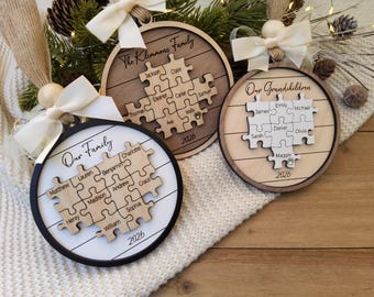May include: Three circular wooden ornaments featuring puzzle piece designs with engraved names and the year 2026. Each ornament is topped with a cream-colored ribbon bow and a wooden bead. Text includes Our Family, The Klemmens Family, and Our Grandchildren.