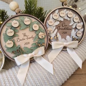 May include: Two circular wooden ornaments featuring layered wreath designs. The left ornament displays The Baxter Family 2026 with names on small baubles. The right ornament shows Our Grandchildren 2026 with names. Both have cream ribbons.