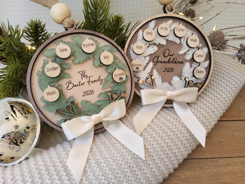 May include: Two circular wooden ornaments featuring layered wreath designs. The left ornament displays The Baxter Family 2026 with names on small baubles. The right ornament shows Our Grandchildren 2026 with names. Both have cream ribbons.