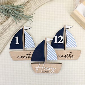 May include: Wooden sailboat-shaped milestone markers. The sailboats have navy blue sails with white and light blue stripes, and white flags. One marker reads "1 month", another "12 months", and the third has the name "Henry James".
