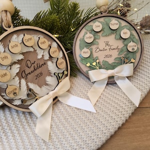 May include: Two circular wooden ornaments featuring a layered wreath design with small wooden tags engraved with names. One ornament reads Our Grandchildren 2026 and the other The Baxter Family 2026. Each piece is finished with a cream satin ribbon bow.