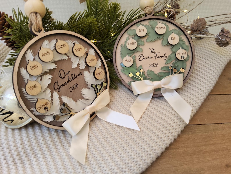 May include: Two circular wooden ornaments featuring a layered wreath design with small wooden tags engraved with names. One ornament reads Our Grandchildren 2026 and the other The Baxter Family 2026. Each piece is finished with a cream satin ribbon bow.