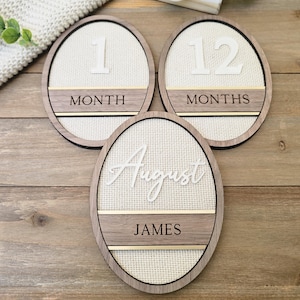 Pode incluir: Três placas ovais de madeira para marcos com um fundo de tecido cor de creme. Duas placas exibem os números 1 e 12 com o texto MONTH ou MONTHS. A terceira placa apresenta o texto em cursivo August e o nome JAMES em letras de forma.