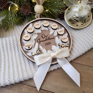 May include: A circular wooden ornament featuring a white feather wreath design, the text "Our Grandchildren 2026," and several small wooden discs engraved with names including Emily, Andy, Carter, Madi, Violet, and Winnie with a paw print icon.