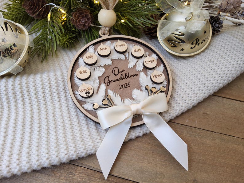 May include: A circular wooden ornament featuring a white feather wreath design, the text "Our Grandchildren 2026," and several small wooden discs engraved with names including Emily, Andy, Carter, Madi, Violet, and Winnie with a paw print icon.