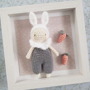 Może przedstawiać: Szydełkowy królik w białym kapeluszu i szarym kombinezonie, stojący w białej ramie na różowym tle. Dwie pomarańczowe szydełkowe marchewki znajdują się na tle.