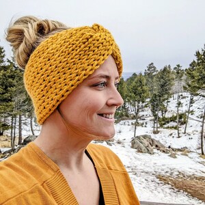 May include: A woman wearing a yellow knitted headband with a knotted design. The headband is thick and warm, perfect for cold weather. The woman is smiling and looking to the side, with a snowy landscape in the background.