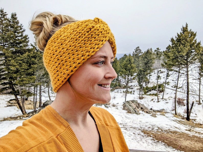 May include: A woman wearing a yellow knitted headband with a knotted design. The headband is thick and warm, perfect for cold weather. The woman is smiling and looking to the side, with a snowy landscape in the background.