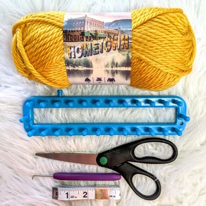 May include: A yellow ball of yarn labeled "Hometown Linen Brand Yarn" with a blue knitting loom, purple crochet hook, scissors, and a measuring tape.