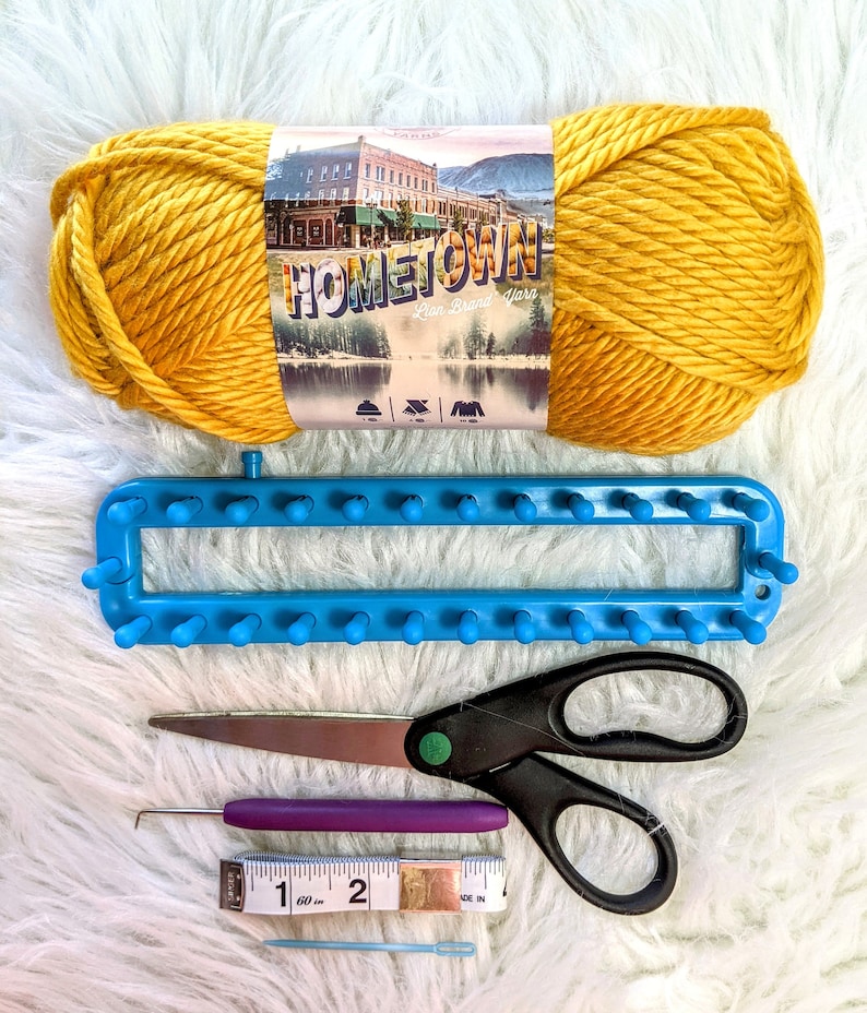 May include: A yellow ball of yarn labeled "Hometown Linen Brand Yarn" with a blue knitting loom, purple crochet hook, scissors, and a measuring tape.
