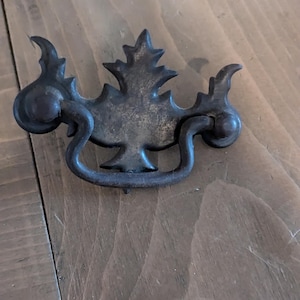 May include: A vintage, ornate, metal drawer pull with a leaf design. The pull is dark brown and has a worn, rustic look.