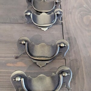 May include: Five antique brass cabinet pulls with a decorative, ornate design. The pulls have a curved, D-shaped handle and a rectangular plate with a scalloped edge.