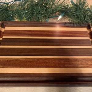 May include: A rectangular wooden cutting board with a natural wood finish. The board is made of multiple strips of wood in different colors, including brown, red, and light wood. The board has a raised edge around the perimeter.