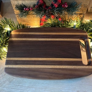May include: A small, oval-shaped wooden cutting board with a handle. The board is made of dark walnut wood with light wood strips running across the grain. The board is sitting on a light wood surface.