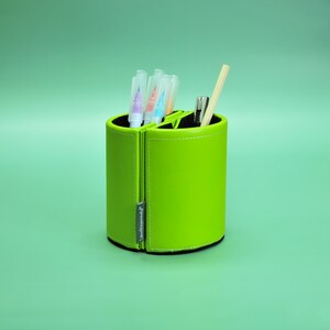 Può includere: Un organizer da scrivania cilindrico verde brillante con interno nero. Contiene penne, matite e altri articoli di cancelleria. L'organizer è diviso in sezioni e ha una chiusura a cerniera. Una piccola etichetta recita "theLegend".