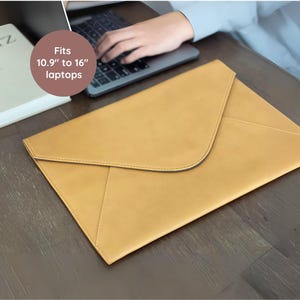 May include: A tan leather laptop sleeve with a curved flap closure. The sleeve is designed to fit laptops from 10.9 to 16 inches. The sleeve is on a dark wooden surface.