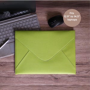 Puede incluir: Una funda para portátil verde lima con cierre de solapa, diseñada para portátiles de 32,8 cm a 36,8 cm. La funda está sobre una superficie de madera, junto a un portátil, un lápiz óptico y un ratón negro.