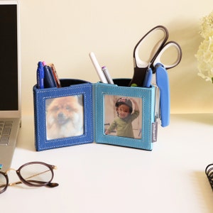 Peut inclure: Un organiseur de bureau bleu et bleu clair avec deux cadres photo. L'organisateur contient des stylos, des ciseaux et une agrafeuse. Un cadre affiche un chien, l'autre un enfant. L'organisateur est sur un bureau blanc.
