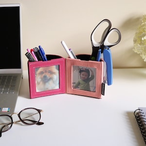 May include: A pink and light pink desk tidy with two photo frames, holding a photo of a dog and a child. The tidy holds pens, scissors, and a stapler. The tidy is on a white desk.