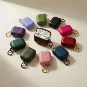 May include: A collection of colorful AirPod cases in various colors, including purple, green, pink, and blue. Each case has a textured finish and a gold or black keyring. One case is open, revealing white earbuds.