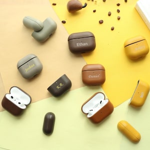 Op de afbeelding: Een collectie van beschermende hoesjes in lederlook voor draadloze oordopjes in diverse kleuren, waaronder bruin, beige, olijfgroen en geel. Verschillende hoesjes zijn gepersonaliseerd met namen of initialen zoals Ethan, Joshua, Daniel, K.R. en MP.