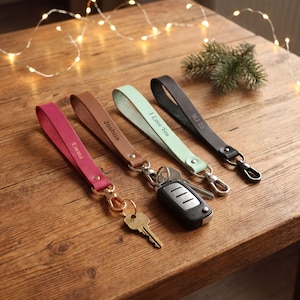 May include: Four leather wristlet keychains in magenta, brown, mint green, and black. Each strap features a metal rivet, a swivel lobster clasp, and custom engraving: Emma, Joshua, I Love You, and MJ 23. One keychain holds a car key fob and a metal key.