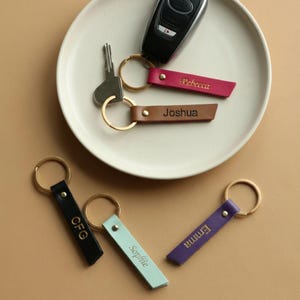 May include: A selection of personalised leather keyrings in various colours: black, teal, purple, brown, and pink. Each keyring has a gold-coloured ring and is engraved with a name or initials. The keyrings are arranged on a white plate and a neutral surface.