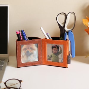 May include: A dual pen holder with photo frames, one brown and one orange, each holding a picture. The brown holder has a picture of a dog, and the orange holder has a picture of a child. Pens, scissors, and a stapler are inside.