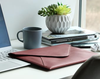 May include: A burgundy leather envelope-style document holder rests on a white desk next to a gray mug and a laptop. A small white decorative pot with a succulent plant sits on a stack of magazines near a window.