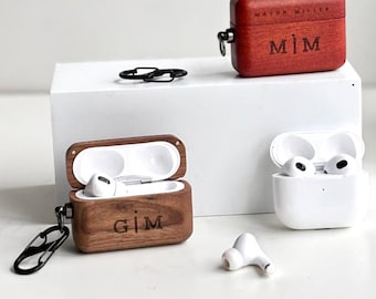 May include: Several rectangular wooden cases for wireless earbuds, featuring engraved names like Renee JONES, M|M, G|M, and Daddo Papi. Some cases include black metal carabiner clips, while others are shown open with white earbuds inside.