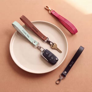 May include: Four leather wristlet keychains in mint green, brown, pink, and black. The mint green strap is engraved with "I Love You" and attached to a car key fob. The brown strap is engraved with "Joshua" and a key. "Emma" is on the pink strap, and "MJ 23" is on the black strap.