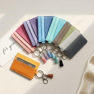 May include: A selection of vibrant cardholders with keyrings and tassels. The cardholders are in shades of pink, purple, blue, and grey. One cardholder displays the text "DIVIDEND" and "CIBC". The keyrings are adorned with tassels in complementary colours.