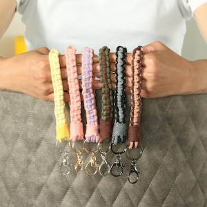 May include: Six macrame wrist keychains in a range of colours: yellow, pink, purple, green, grey, and brown. Each keychain has a metal clasp for attaching keys or small items. The keychains are displayed against a grey background.