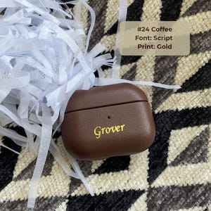 Dark brown personalized leather AirPods case with custom monogram in script font and gold heat stamping, tech accessory, minimalist aesthetic protective earbud case.