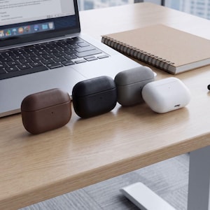 May include: Four wireless earbud charging cases arranged in a row. The cases are brown, black, gray, and white. Each case has a smooth, rounded rectangular shape with a small indicator light on the front and a charging port visible on the white case.