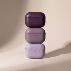 May include: A vertical stack of three rectangular earbud charging cases with rounded corners. Each case features a textured finish in a different shade of purple, ranging from a deep dark plum at the top to a medium mauve in the middle and a light lavender at the base.
