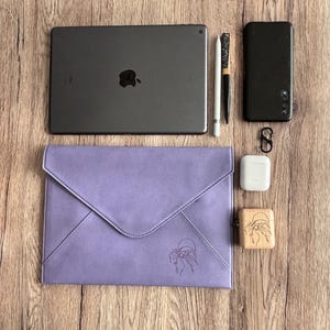 Può includere: Una collezione di accessori tecnologici e una custodia lilla a forma di busta. Gli articoli includono un tablet, un telefono, uno stilo, una penna, auricolari wireless, una custodia per auricolari in legno e un moschettone nero. La custodia presenta un design inciso.