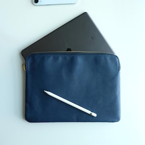 May include: A navy blue leather tablet sleeve with a gold zip, holding a tablet. A white stylus pen rests on the sleeve. A light blue smartphone is visible in the background. The sleeve is designed to protect and carry tablets.