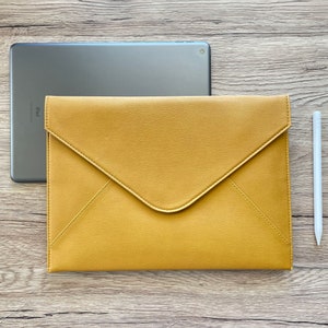 May include: A mustard-yellow envelope-style tablet case with a matching stylus and a silver tablet. The case is made of a smooth material and has a flap closure. The tablet is partially visible behind the case.