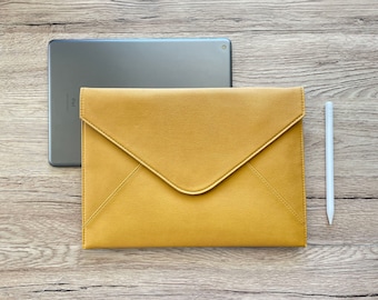 May include: A mustard-yellow tablet case in an envelope style, with a matching stylus and a silver tablet. The case is made of a smooth material and has a flap closure. The tablet is partially visible behind the case.