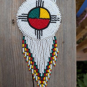 Peut inclure: Un collier médaillon circulaire en perles avec un fond blanc et un motif central à quatre quadrants en vert, jaune et rouge, accentué par des lignes noires. La pièce comprend une longue frange de perles blanches avec des accents rouge, jaune et vert.