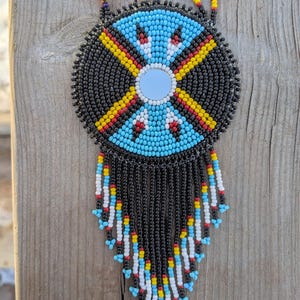 May include: A beaded necklace with a circular pendant featuring a central mirror and a geometric design in black, blue, yellow, and red. The pendant has a beaded fringe in various colors. The necklace is displayed on a wooden surface.
