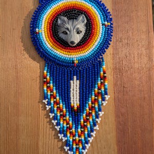 May include: A beaded necklace featuring a wolf's face in the centre. The necklace is made from blue, white, red, yellow and black beads. The wolf's face is grey and white.