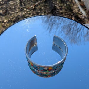May include: A silver cuff bracelet with a beaded design. The bracelet has a band of turquoise, yellow, red, and white beads. The bracelet is open at the top and is displayed on a reflective surface.