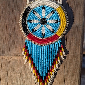 May include: A beaded necklace with a circular pendant. The pendant has a central starburst design in white and black, surrounded by blue, yellow, black, and red beads. The necklace has a beaded fringe in blue, red, yellow, and black.
