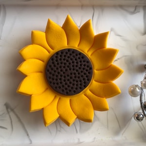 May include: A yellow felt sunflower with a brown felt center. The sunflower is on a white marble surface.
