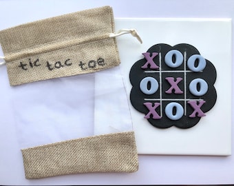Könnte beinhalten: Ein Jutesack mit einem durchsichtigen Plastikfenster und dem aufgedruckten Text "tic tac toe". Der Sack liegt neben einem schwarzen Tic-Tac-Toe-Brett mit violetten X und blauen O.